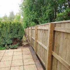 Wooden fence along a garden path with trees and shrubs on the left. The path is flanked by square stone tiles bordered by a brick line on the right.