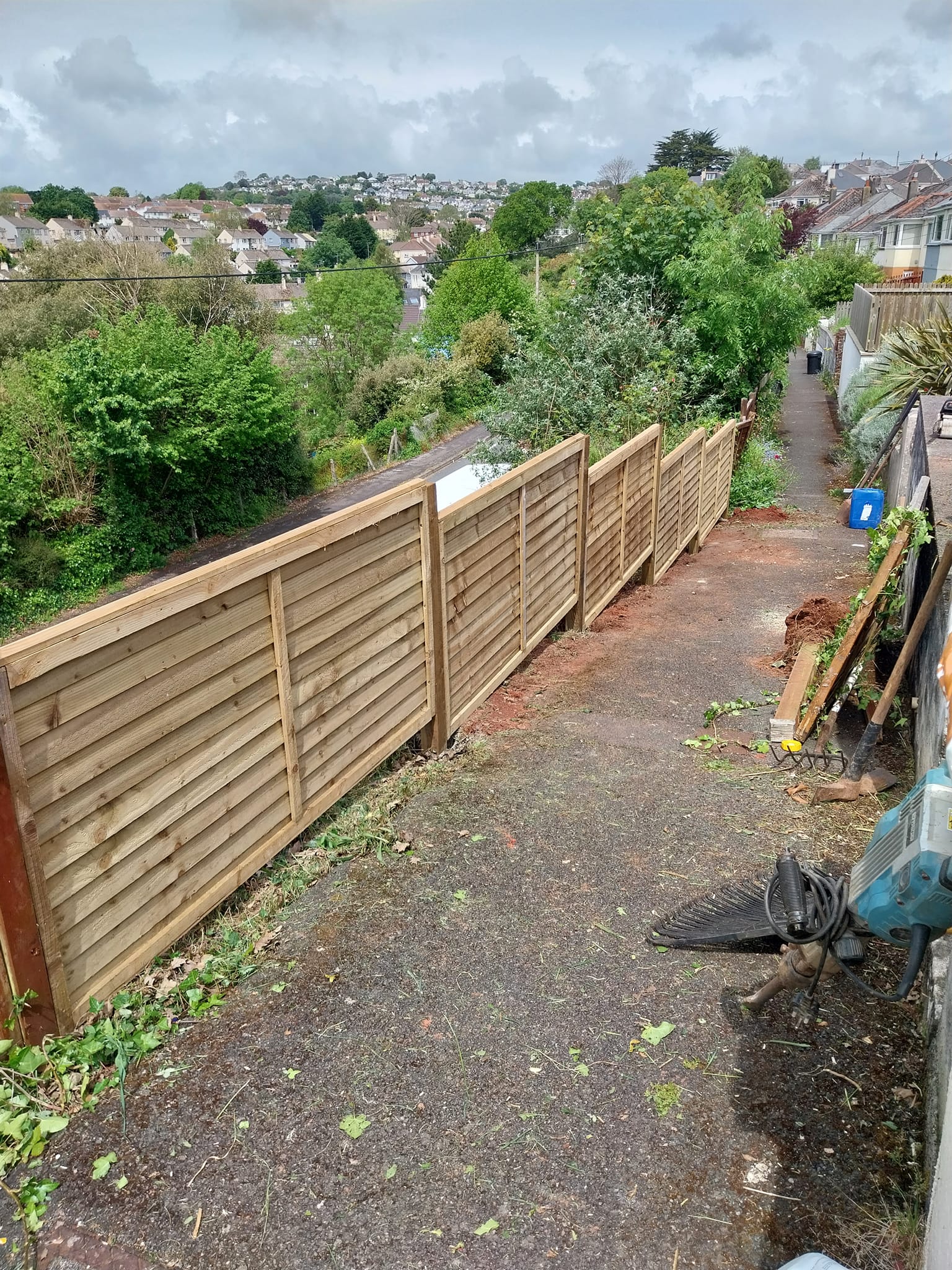 Newly installed wooden fence along a narrow, paved path with greenery on both sides. The path is slightly sloped, and construction materials are scattered on the ground. Residential houses are visible in the background under a cloudy sky.