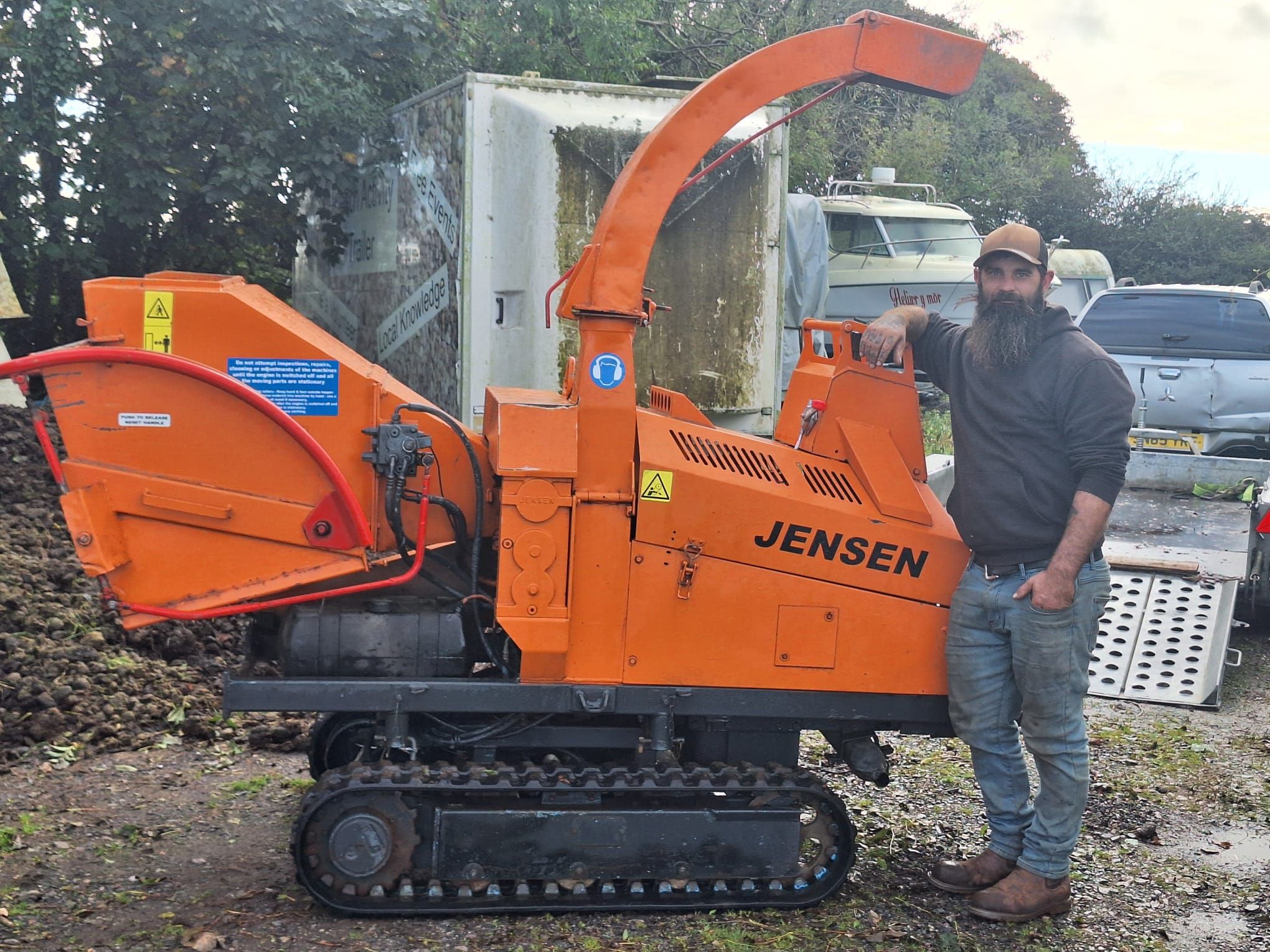 A man with a beard and cap stands next to an orange Jensen wood chipper with tracks. The machine has a long chute and warning stickers. In the background, there are trees, a vehicle, and a white container.
