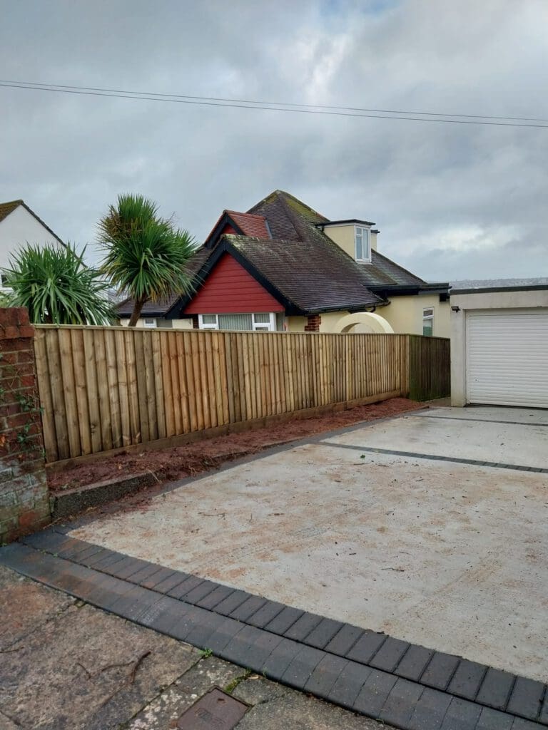 A house with a red gable roof and beige walls is partially visible behind a tall wooden fence. A small palm tree stands in front. In the foreground, there is a freshly paved driveway. The sky is overcast with clouds.