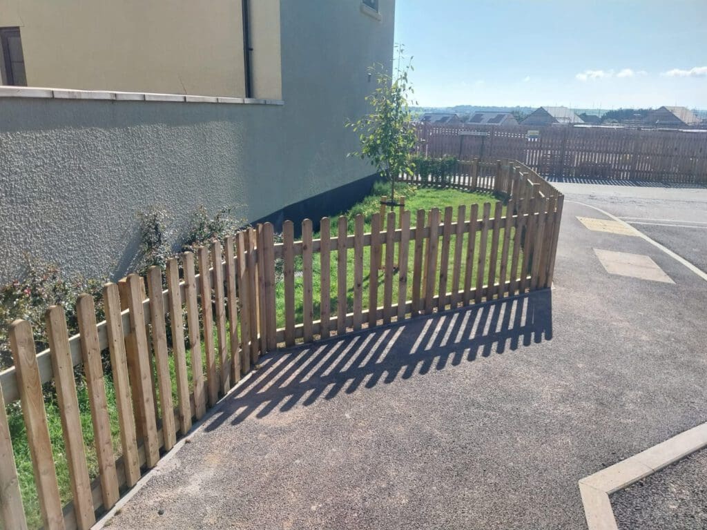 A small, enclosed grassy area with a wooden picket fence near a building. There's a young tree in the middle of the grass. The pavement beside it leads towards a road under a clear, blue sky.