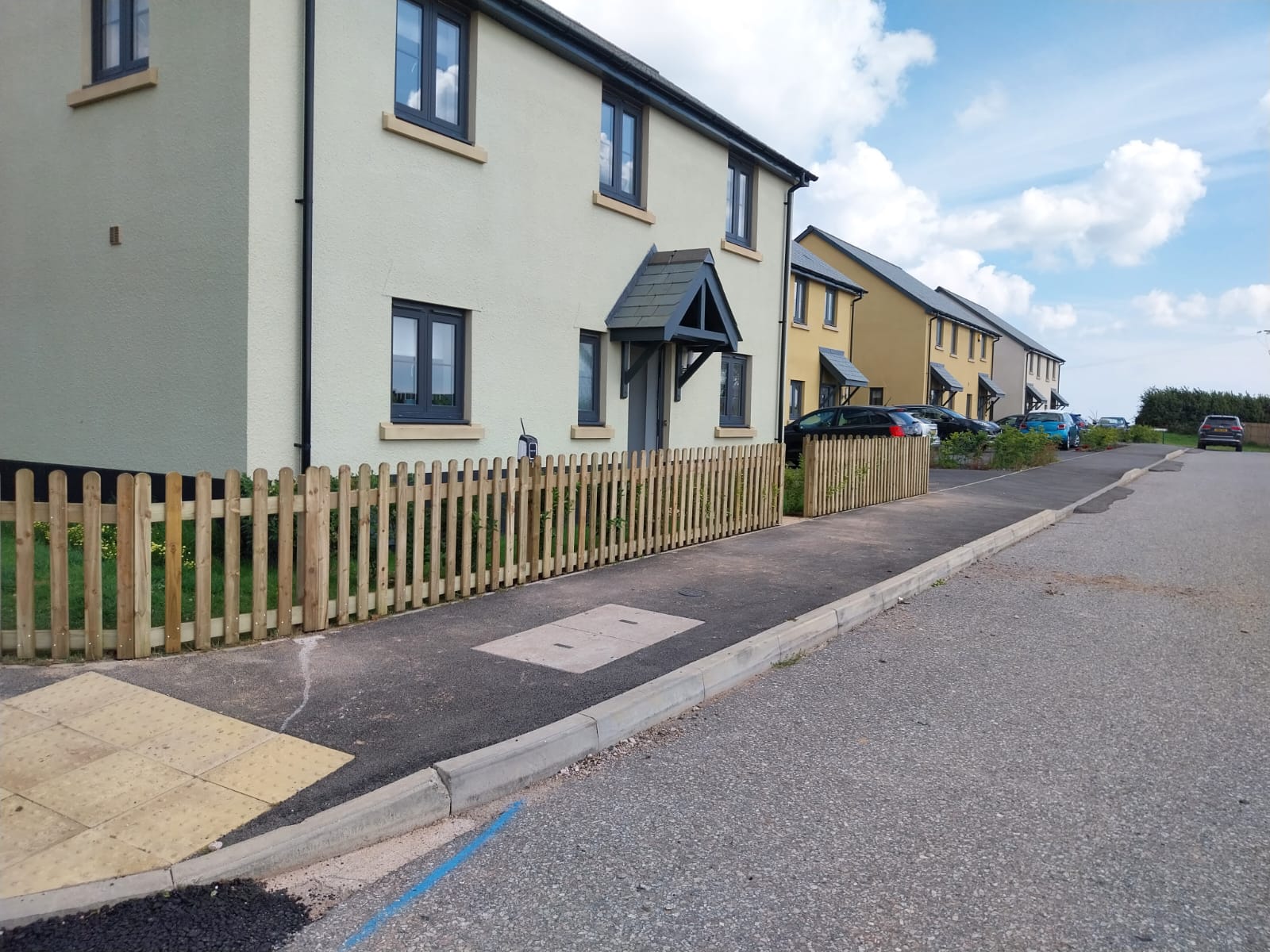 Suburban street with modern two-story houses in beige and green. Wooden fencing lines the walkway. A blue sky with fluffy white clouds is above. The road curves, bordered by grass and pavement.