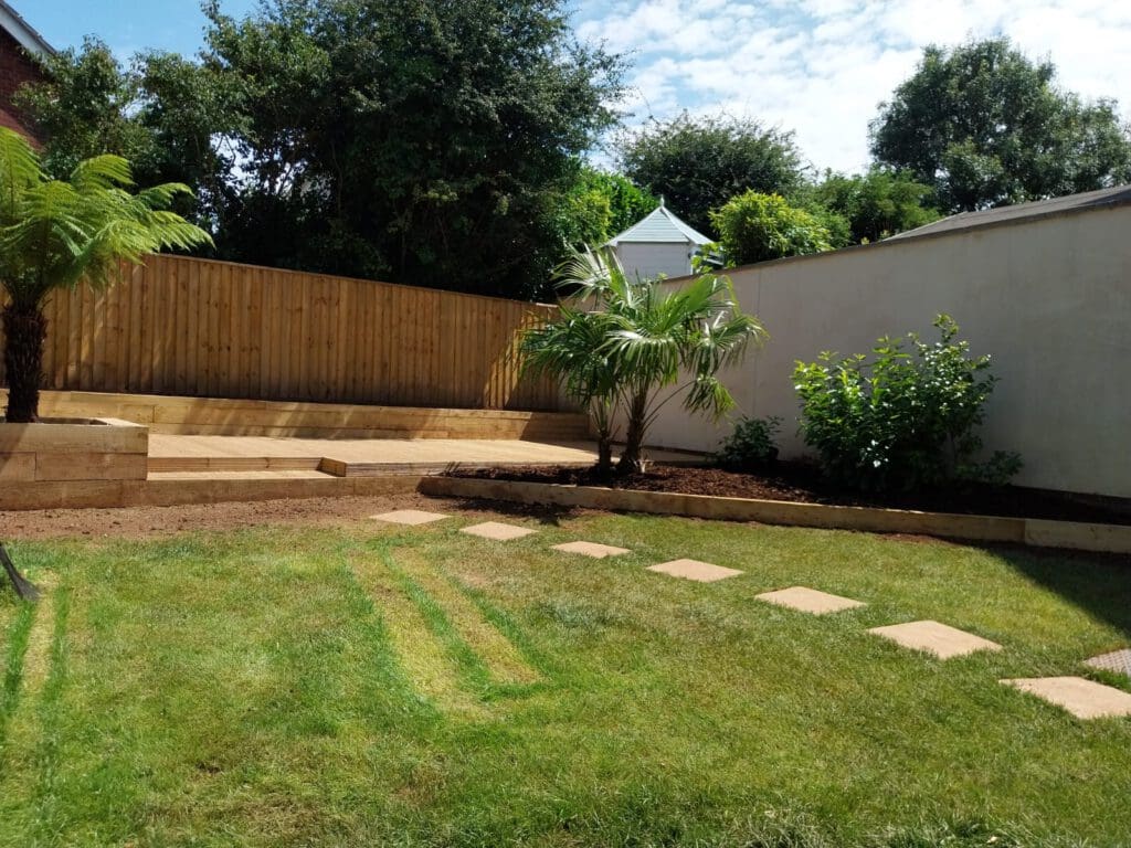 A backyard garden featuring a lush green lawn with stepping stones leading to a raised wooden deck. Tall trees and a small white shed are in the background. A palm-like plant and shrubs are beside the deck, under a partly cloudy sky.