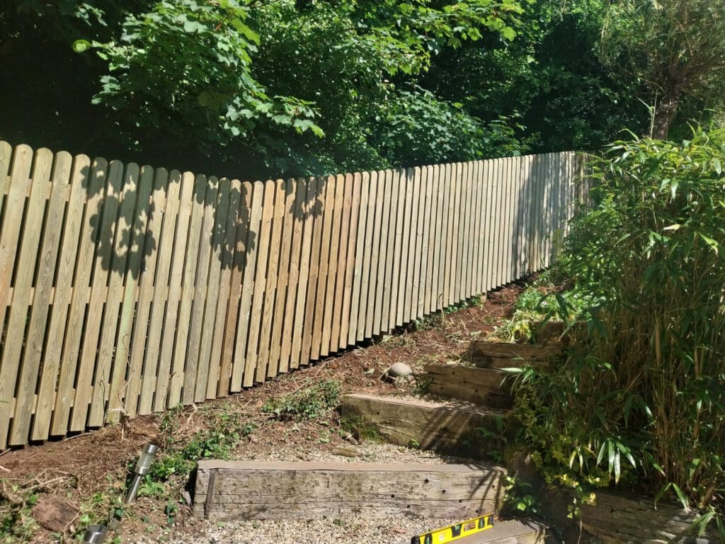 A tall wooden fence runs along a garden with lush greenery. The ground is uneven, and wooden steps lead up the slope. Sunlight filters through the trees, casting shadows on the fence. A yellow level lies on the ground.
