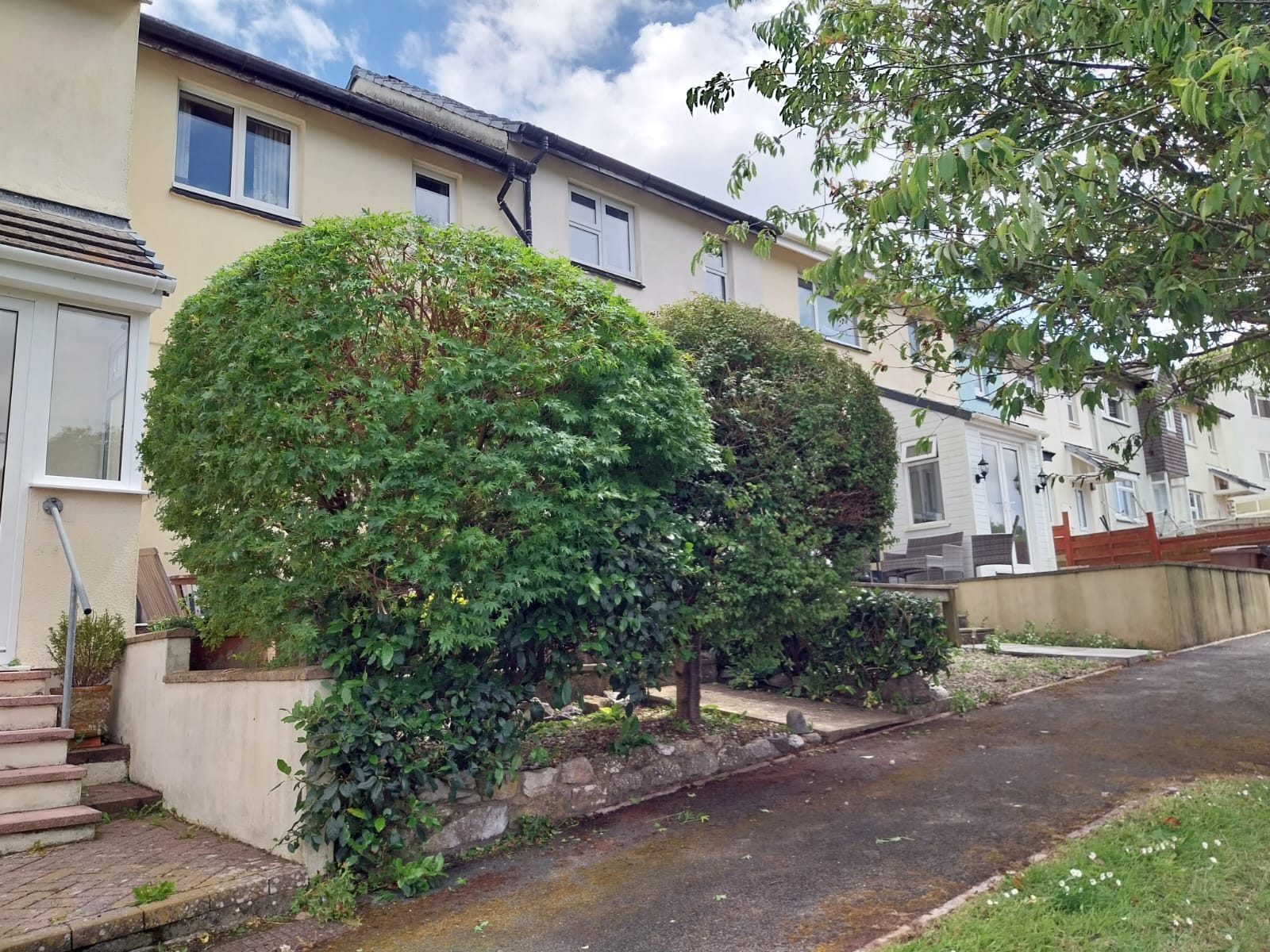 A row of two-story houses with cream and white exteriors. Prominent round bushes flank a path leading to one house. The scene includes a paved walkway, stairs, and a foreground tree with green leaves under a partly cloudy sky.