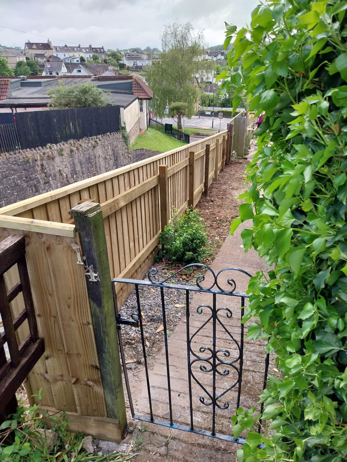 View of a narrow pathway with a metal gate and wooden fence on one side, lined with greenery. The path leads downhill towards a residential area with houses and a street visible in the background under a cloudy sky.