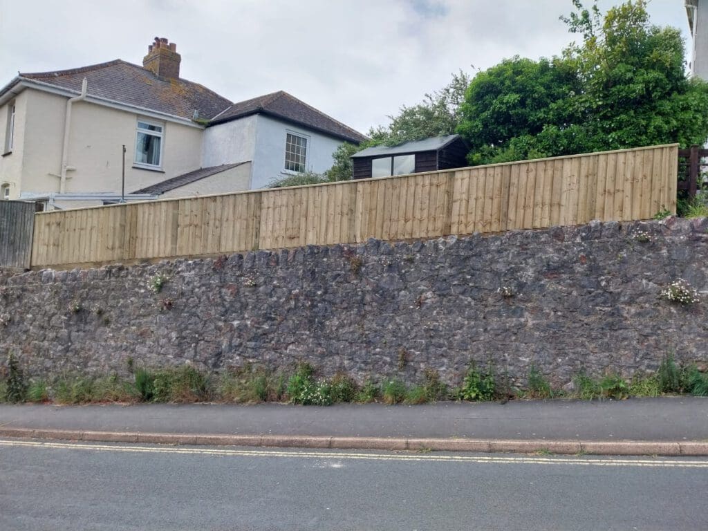 A stone retaining wall along a roadside with a wooden fence on top. Behind the fence, a house with a sloped roof is partially visible. There are shrubs and plants growing on the wall and a small structure visible behind the fence.