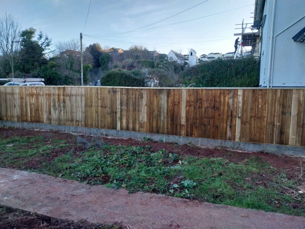 A newly built wooden fence enclosing a yard with fresh soil and sparse greenery. The background features a residential area with houses, a tree-lined horizon, and a person on scaffolding working on a building.