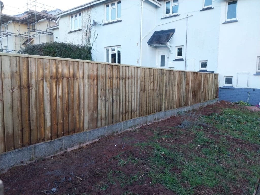A wooden fence separates a garden from a white two-story house. The garden has patches of grass and soil. The house has a porch and is partially covered in scaffolding on the left side.