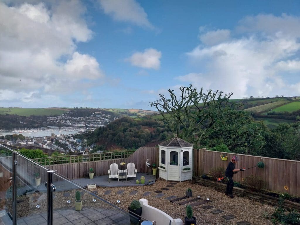 A person playing croquet in a garden with a view of rolling hills, a town, and a river. The garden has a small gazebo, potted plants, and a glass railing. The sky is partly cloudy, and the scene is tranquil and scenic.
