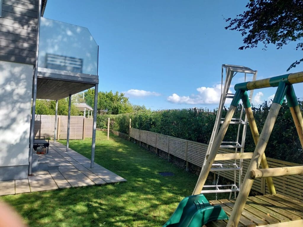 A backyard with a modern house on the left, featuring a patio and a glass balcony. A ladder leans against a wooden swing set on the right. A lawn and hedges extend into the distance under a clear blue sky.
