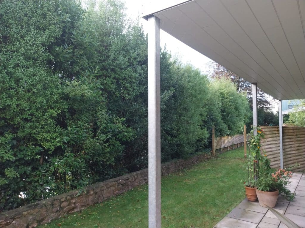 A covered patio with a slanted roof overlooks a garden. The patio has a metal pole and the ground is paved with tiles. The garden features a stone path, green grass, dense hedges, and potted plants on the patio. A wooden fence runs along the hedge.