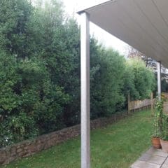 A covered patio with a slanted roof overlooks a garden. The patio has a metal pole and the ground is paved with tiles. The garden features a stone path, green grass, dense hedges, and potted plants on the patio. A wooden fence runs along the hedge.