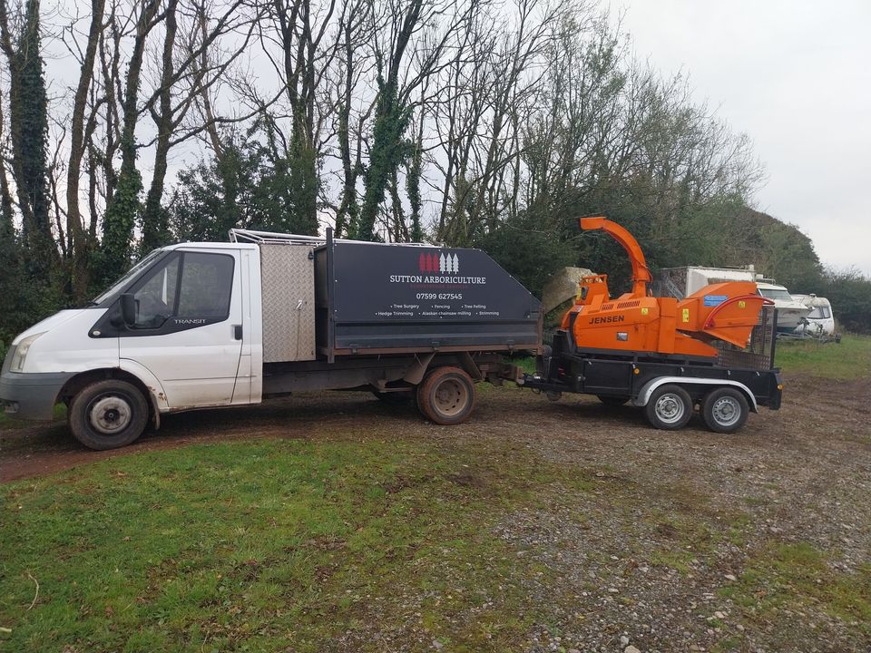 A white truck with a Sutton Arboriculture logo is parked on grass near trees. It is towing an orange wood chipper. There are more vehicles in the background amidst greenery.